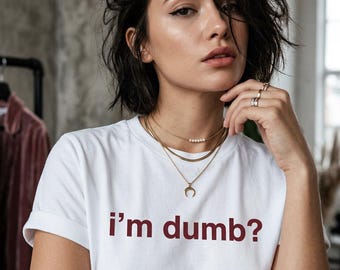 Puede incluir: Una camiseta blanca de manga corta con el texto "i'm dumb?" impreso en una fuente sans-serif de color rojo oscuro en el pecho. La prenda tiene un cuello redondo clásico y se lleva con collares dorados superpuestos y varios anillos.