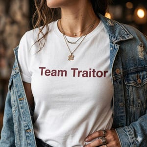 Puede incluir: Camiseta blanca con el texto "Team Traitor" en color granate. La camiseta se lleva con una chaqueta vaquera y collares dorados. La imagen es un plano medio, centrado en la camiseta y la parte superior del cuerpo.