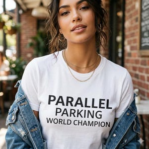 Może przedstawiać: Biały t-shirt z krótkim rękawem z czarnym napisem PARALLEL PARKING WORLD CHAMPION nadrukowanym z przodu. Koszulka jest noszona pod niebieską kurtką dżinsową. Wzór jest wyśrodkowany na klatce piersiowej i wykonany pogrubioną czcionką bezszeryfową.
