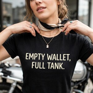 Könnte beinhalten: Ein schwarzes Kurzarm-T-Shirt mit dem Text "EMPTY WALLET, FULL TANK." in einer hellen serifenlosen Schrift. Das Shirt wird mit einem schwarz-beigen Halstuch und einer goldfarbenen Halskette mit einem kleinen Zapfsäulen-Anhänger kombiniert.