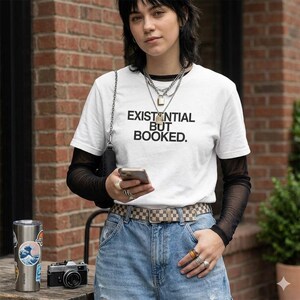 Może przedstawiać: Biała koszulka z napisem "EXISTENTIAL BUT BOOKED". Osoba ma na sobie czarną koszulkę z długim rękawem z siatki. Osoba ma na sobie kraciasty pasek i niebieskie dżinsy. Kubek ze stali nierdzewnej i aparat fotograficzny znajdują się na drewnianej powierzchni.