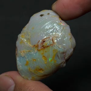 Op de afbeelding: Een ruwe, ongeslepen opaal edelsteen. De steen heeft een bleke, melkachtige witte basiskleur met blauwe, gele en bruine gebieden. Het oppervlak heeft een textuur met zichtbare lagen en insluitsels. De opaal wordt tussen twee vingers gehouden.
