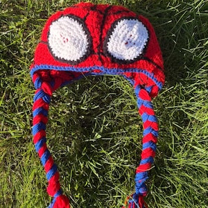 Puede incluir: Un gorro de ganchillo rojo y azul con detalles de ojos blancos, que se asemeja a una máscara de superhéroe. El gorro presenta orejeras trenzadas en rojo y azul, con borlas rojas en los extremos. El gorro está diseñado para ser usado en la cabeza.