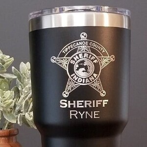 May include: Black Yeti tumbler with a silver lid and base. It is engraved with a sheriff's star and the text "Tippecanoe County Indiana Sheriff Ryne".