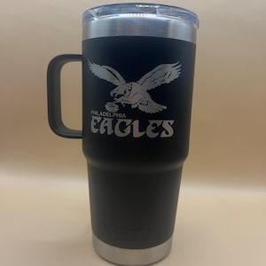 May include: Black stainless steel travel mug with a black handle. The mug has a white eagle graphic with the words "Philadelphia Eagles" underneath.