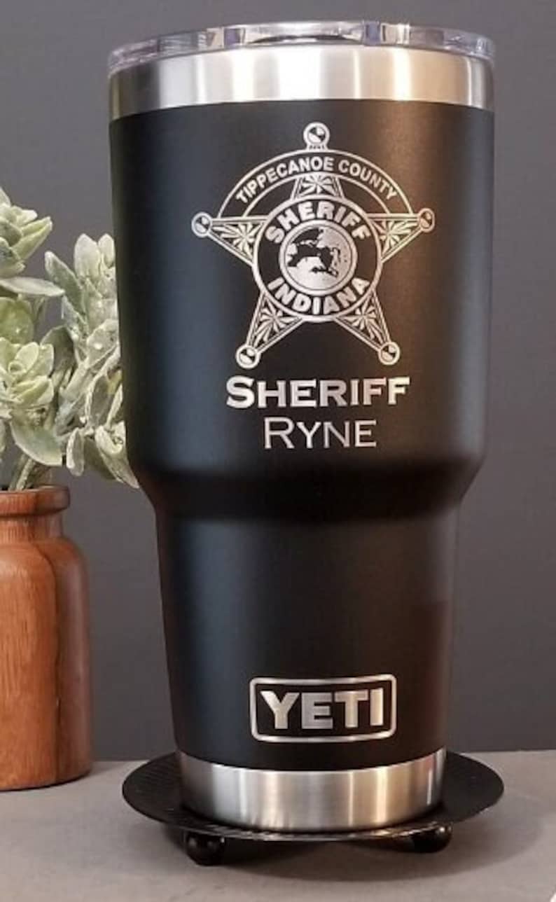 May include: Black Yeti tumbler with a silver lid and base. It is engraved with a sheriff's star and the text "Tippecanoe County Indiana Sheriff Ryne".