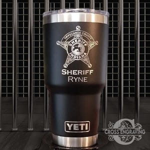 May include: Black Yeti tumbler with a silver lid and base. The tumbler is engraved with a sheriff's star badge, the words "Tippecanoe County Sheriff Indiana", and the name "Sheriff Ryne".