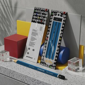 Puede incluir: Un lápiz azul Staedtler Mars Lumograph 2B yace horizontalmente frente a dos tarjetas de embalaje rectangulares verticales. Las tarjetas presentan un patrón repetitivo en blanco y negro del logotipo printland y una ilustración gráfica de un lápiz azul.