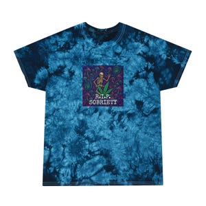 May include: A blue tie-dye t-shirt with a square graphic featuring a skeleton, cannabis leaf, and the text "R.I.P. SOBRIETY." The graphic has a psychedelic design with vibrant colors. The shirt is made of a soft material.