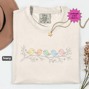 Può includere: T-shirt color avorio con scollo tondo, con un ramo con sei uccelli dai colori pastello. La maglietta ha un'etichetta con la scritta "Comfort Colors". Un pendente a stella dorata pende da una catena d'oro. Il testo "multiple color choices" è visibile.