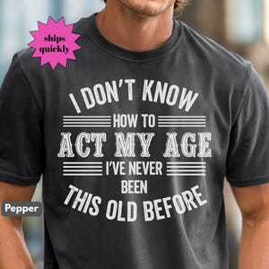 May include: A dark gray t-shirt with the text "I DON'T KNOW HOW TO ACT MY AGE I'VE NEVER BEEN THIS OLD BEFORE" in white. The shirt has a crew neck and short sleeves. A pink starburst graphic says "ships quickly".