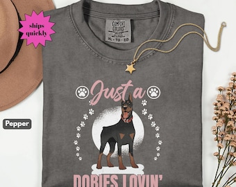 May include: A gray Comfort Colors t-shirt featuring a Doberman Pinscher graphic with the text "Just a Dobies Lovin'". The design includes paw prints and a pink banner. A gold star necklace is draped on the shirt.