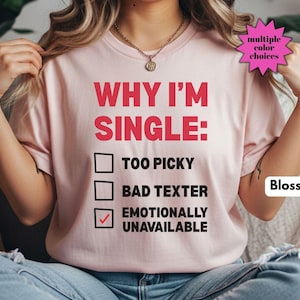 May include: A light pink t-shirt with the text "WHY I'M SINGLE:" in red, followed by three check boxes. The bottom box is checked. The shirt also has the word "Blossom" in a white box and a pink starburst that says "multiple color choices".