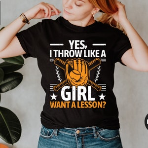 May include: Black t-shirt with the text "YES, I THROW LIKE A GIRL WANT A LESSON?" in white and orange. The design includes a baseball glove, baseball, and crossed bats. The shirt is a crew neck.