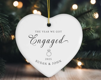 May include: A white heart-shaped ceramic ornament hanging from a gold string. The text reads THE YEAR WE GOT Engaged, followed by a line drawing of a diamond ring, the year 2025, and the names SUSAN & JOHN in black lettering.
