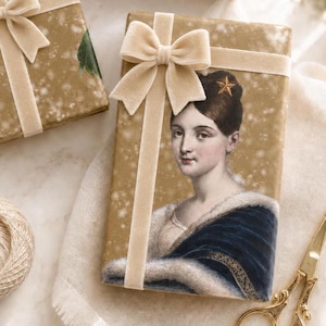 K&ouml;nnte beinhalten: Eine rechteckige Geschenkbox in goldenem Papier mit dem Portr&auml;t einer Person mit dunklem Haar und einem Stern-Accessoire, verziert mit einer beigen Samtschleife. Das Portr&auml;t zeigt eine Person in einem dunkelblauen Kleidungsst&uuml;ck mit Pelzbesatz.