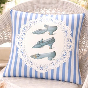 May include: Square throw pillow with a blue and white striped pattern. The center features a white lace-edged circle with illustrations of three pairs of light blue vintage shoes. The pillow is placed on a white wicker chair.