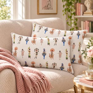 May include: Two rectangular throw pillows with a white background featuring a repeating pattern of illustrated figures in elaborate, colorful costumes, including feathered headpieces, dresses, and patterned outfits in shades of orange, blue, and green.