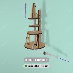 May include: A wooden rocket launcher model with a syringe attached. The model has multiple circular platforms and a rocket-shaped top. Text on the image reads "ROCKET LAUNCHER" and "EASY BUILD - 10 min".