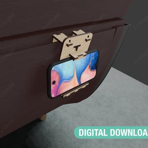 May include: A wooden phone holder attached to a dark brown surface. The holder is designed to hold a smartphone in a vertical position. The phone is visible in the holder, displaying a colorful abstract background.