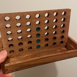 K&ouml;nnte beinhalten: Ein Connect-Four-Spiel aus Holz mit braunem Finish. Das Spielbrett hat eine Reihe kreisf&ouml;rmiger L&ouml;cher zum Einwerfen von Spielsteinen. Vier gr&uuml;ne Spielsteine sind auf dem Brett sichtbar. Das Spiel steht auf einem Holzsockel.
