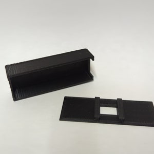 May include: A black plastic box with a removable lid. The lid has two tabs on the side and a rectangular opening in the center.