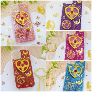 May include: A collection of glittery phone cases featuring Sailor Moon-inspired motifs, including heart-shaped compacts, star emblems, and crescent moon wands in various colours like pink, purple, red, and blue, with gold-toned decorative accents.