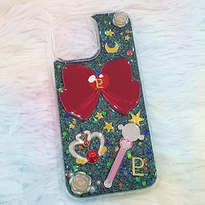May include: Clear phone case with a glittery green background, adorned with a large red bow, stars, a crescent moon, and a pink wand. The case features a gold letter "P" and various decorative elements.