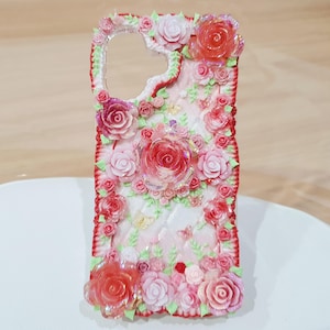 Puede incluir: Una funda de teléfono rectangular con un fondo rosa claro acolchado, decorada con numerosas rosas de resina tridimensionales en tonos rojo, rosa y blanco. Pequeñas hojas verdes y diminutas mariposas amarillas se intercalan entre las flores.
