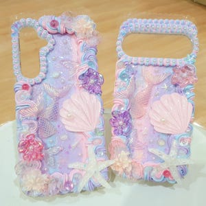 May include: Two rectangular phone cases decorated with pastel-coloured whipped cream texture, featuring iridescent mermaid tails, pink seashells, white starfish, and translucent purple and pink floral accents with small pearl beads throughout.