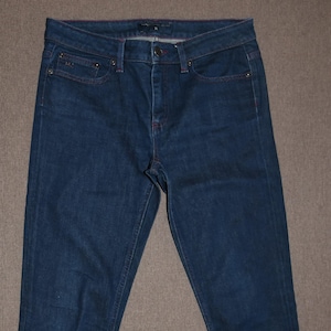 Może przedstawiać: Jeans z ciemnoniebieskiego denimu o prostym kroju nogawek. Jeans mają zapięcie na guziki i szlufki na pasek.