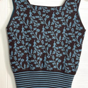 Peut inclure: Débardeur tricoté marron et bleu avec un motif floral. Le haut présente une encolure carrée et un ourlet rayé. Le motif floral est bleu clair sur fond marron. Le débardeur est suspendu à un cintre en bois.
