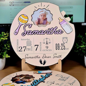 May include: A personalized wooden baby announcement plaque with the name "Samantha" in cursive. The plaque includes a photo, birth date, time, weight (7 pounds, 9 ounces), and other baby-related icons. The plaque is displayed on a wooden stand.