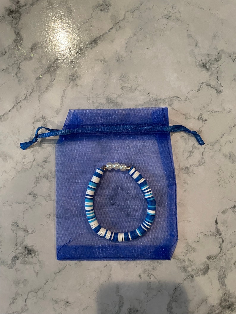 May include: A blue and white beaded bracelet with white pearl accents, displayed in a blue organza bag.