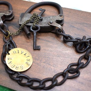 Puede incluir: Grilletes metálicos antiguos con dos cerraduras, llaves y una cadena. El metal tiene un aspecto envejecido y desgastado. Una etiqueta circular dice "GREAT BRITAIN". Los objetos están sobre una superficie de madera.