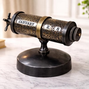 May include: A decorative perpetual calendar with a dark bronze finish. The cylindrical body features an ornate floral pattern and displays "JANUARY 28". The calendar sits on a round base and has a brass-colored ring.