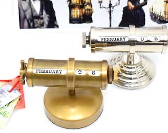 May include: Two cylindrical desk calendars with rotating dials. One is made of brushed brass, the other of polished silver-tone metal. Both display the text FEBRUARY and the number 06. Each features a circular base and side adjustment knobs.