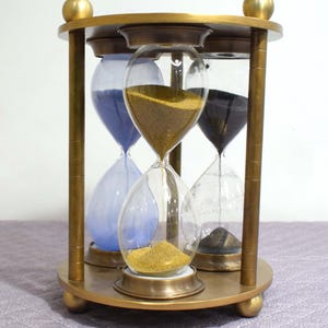 May include: A brass hourglass stand with three separate hourglasses. The hourglasses are filled with blue, gold, and black sand.