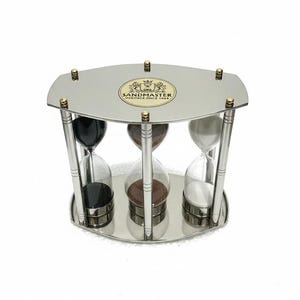 May include: A triple hourglass set featuring a polished silver-tone metal frame with gold-tone accents. The device contains three glass bulbs filled with black, brown, and white sand. A circular logo on the top reads SANDMASTER HERITAGE SINCE 1988.