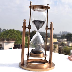 May include: A brass hourglass with a black sand timer. The hourglass is on a white surface.