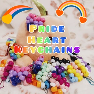 May include: A collection of colorful beaded heart-shaped keychains with rainbow accents. The keychains are made with small plastic beads in various colors, including pink, purple, blue, green, yellow, and orange. The keychains are arranged on a plush toy with the text "PRIDE HEART KEYCHAINS" displayed above them.