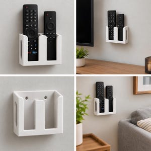 May include: A white wall-mounted remote control holder with two vertical slots. The rectangular organizer features open sides and two circular mounting holes on the back panel, designed to store two black remote controls securely.