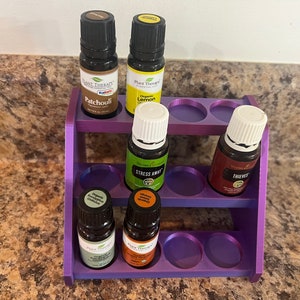 May include: A purple essential oil bottle holder with two shelves, holding several small glass bottles of essential oils. The bottles have black caps and labels with text like "Patchouli", "Lemon", and "Stress Away".