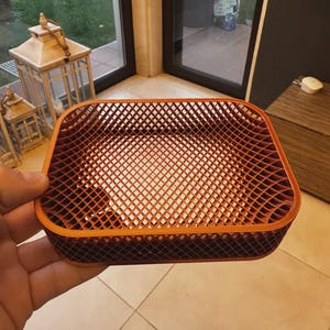 Puede incluir: Una cesta rectangular calada en tonos naranja y marrón. La cesta tiene un diseño de celosía y está sujeta por una mano. Los bordes de la cesta son redondeados y el interior es visible. La cesta se utiliza probablemente para guardar o exhibir.
