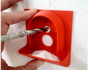 May include: A vibrant orange drill guide and dust catcher is pressed against a white wall. A drill bit is inserted into the guide's hole. The guide has a lower tray filled with white powder, likely collected dust from drilling. The tool is designed to catch dust.