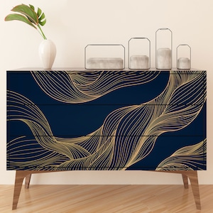 May include: A three-drawer dresser with a navy blue finish and a gold abstract wave pattern. The dresser has wooden legs and is sitting on a wooden floor.