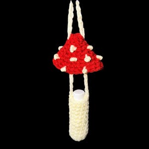 May include: A handmade crochet mushroom-shaped bottle holder. The mushroom cap is red with white spots, and the stem is a cream color. The holder is designed to carry a small bottle, with cream-colored straps for hanging.