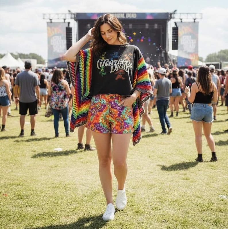 Op de afbeelding: Een vrouw draagt een zwart t-shirt met de tekst "Grateful Dead" en een regenboog gehaakte poncho. Ze draagt ook kleurrijke shorts met patronen en witte sneakers. De achtergrond toont een muziekfestival met een podium en een menigte.
