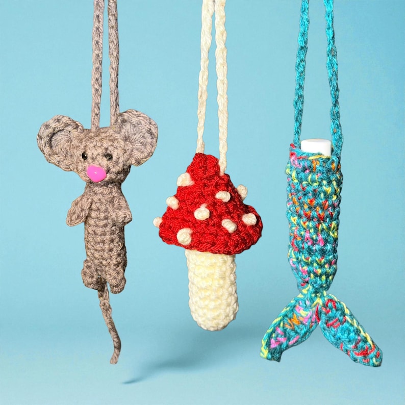 May include: Three handmade crochet ornaments hang against a blue background. A gray mouse with a pink nose, a red mushroom with white spots, and a teal mermaid tail are suspended by cords. These whimsical ornaments are perfect for adding a touch of handmade charm.