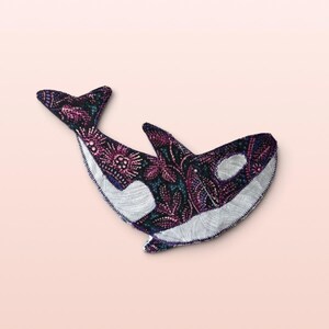 May include: A fabric orca whale ornament featuring a dark purple and black floral pattern with white fabric accents on the eye patch, belly, and dorsal fin area. The edges are finished with a visible dark thread zigzag stitch throughout.